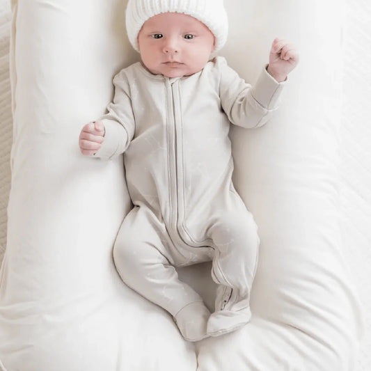 Baby in a grey zipper footie sleeper and knit toque lounges on a cushion