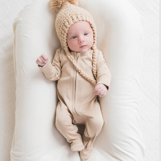 Image of a small baby with blue eyes wearing a warm beige colored sleeper and knit toque lounging on a cushion