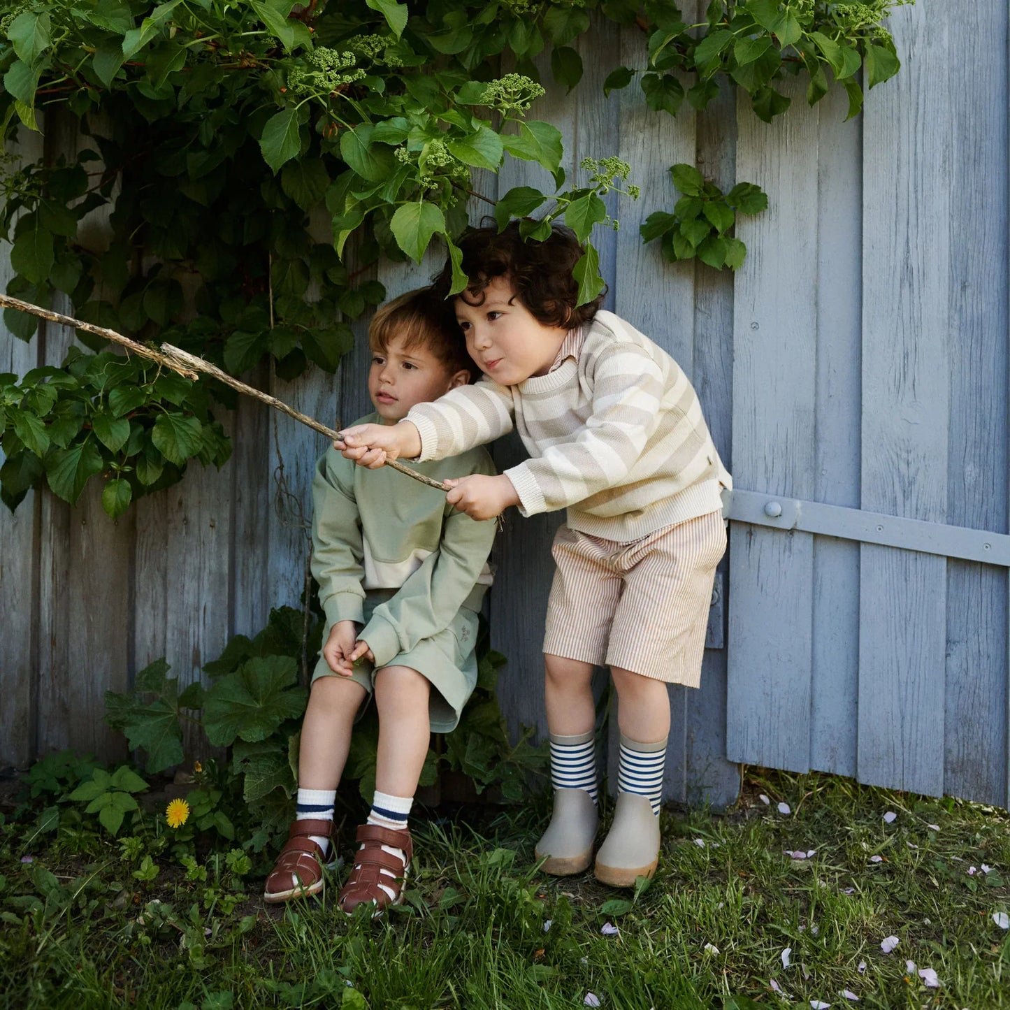 Lifestyle product image of two little boys dressed in shorts, sweaters and rainboots playing in a backyard