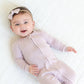 Dark haired baby laying on a muslin wearing a light lilac zip footie sleeper and hair bow