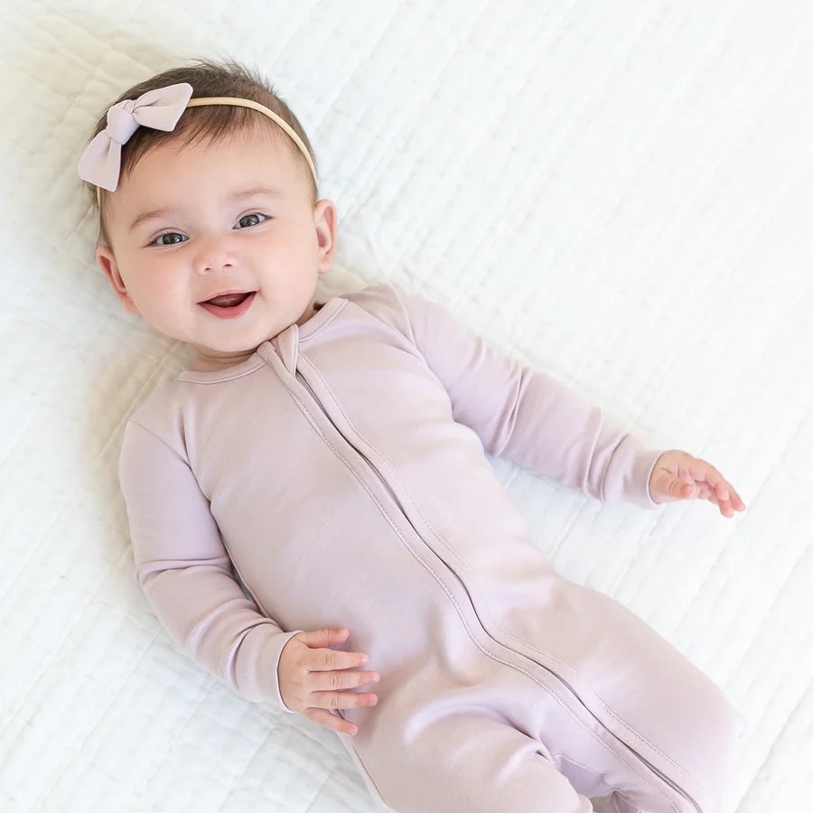 Dark haired baby laying on a muslin wearing a light lilac zip footie sleeper and hair bow